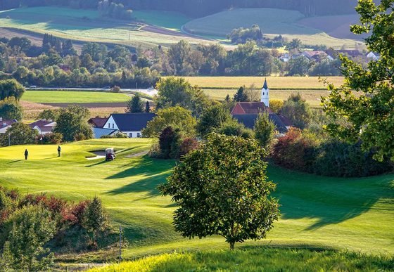 Golfplatz in hügeliger Landschaft mit Golfern, Dorf, Feldern und kleiner Kirche im Hintergrund.