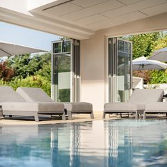 Innenpool mit Liegen, offenen Glastüren, Sonnenschirmen und Gartenblick in einem Wellnessbereich.
