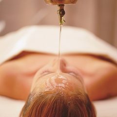 Eine Frau liegt auf einer Liege, während bei einer Wellnessbehandlung Öl auf ihre Stirn tropft.