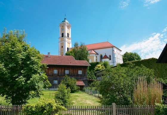 Kirche mit hohem Turm über Garten, Bäumen und Holzhaus in einem Dorf unter blauem Himmel.