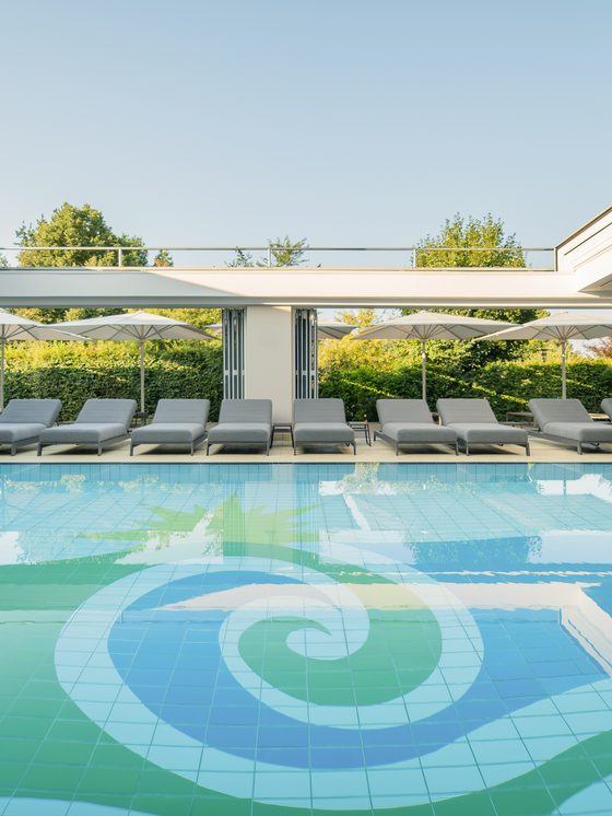 Moderner Innenpool mit Liegen und geöffneten Glasfronten zum begrünten Außenbereich.