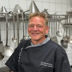 Lächelnder Küchenmeister in schwarzer Kochjacke in einer professionellen Küche vor aufgehängten Kochutensilien.