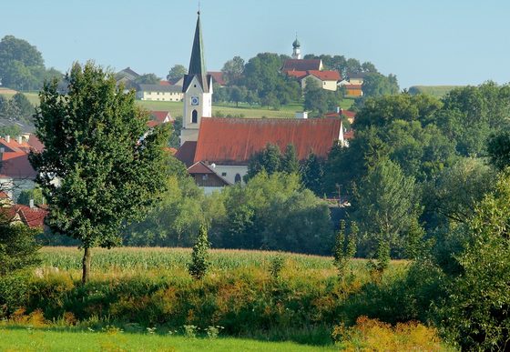 Ein Fluss vor grünen Wiesen und Bäumen mit einem Dorf und einer Kirche im Hintergrund.