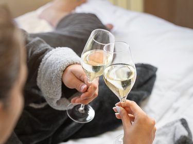 Zwei Personen in Bademänteln stoßen auf einem Bett im Hotelzimmer mit Sektgläsern an.