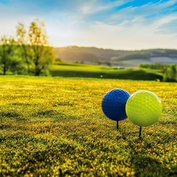 Zwei Golfbälle auf Tees im Gras vor einer grünen Hügellandschaft unter blauem Himmel.