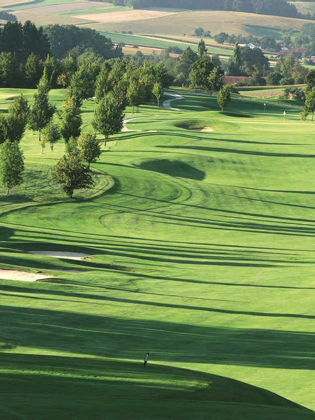 Golfplatz mit gepflegtem Fairway, Bäumen und Sandbunkern in hügeliger Landschaft.