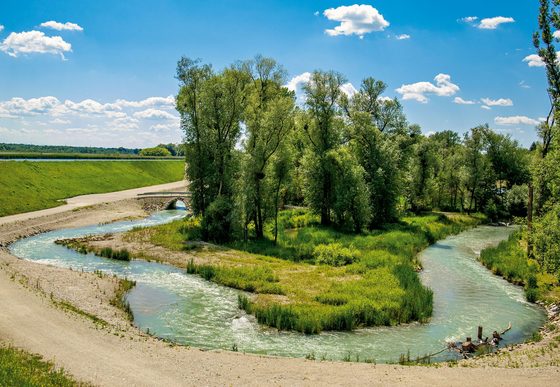 Geschwungener Wasserlauf mit kleiner bewachsener Insel, Bäumen und Kieswegen unter blauem Himmel.