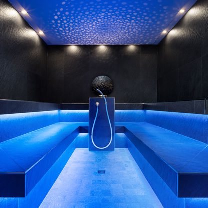 Modernes Dampfbad mit blauer Beleuchtung, schwarzen Wänden, seitlichen Sitzbänken und zentraler Dusche.