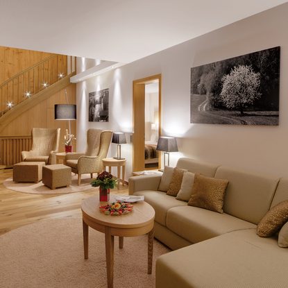 Wohnbereich einer Suite mit Sofa, Sesseln, Couchtisch und Holztreppe im Innenraum.