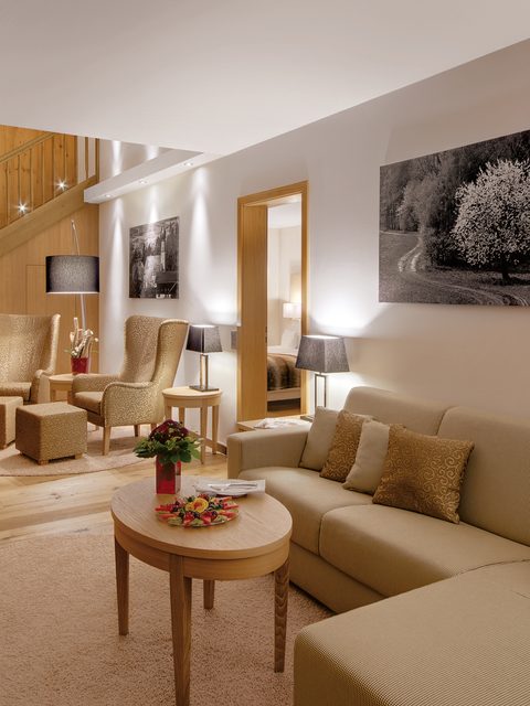 Wohnbereich einer Suite mit Sofa, Sesseln, Couchtisch und Holztreppe im Innenraum.