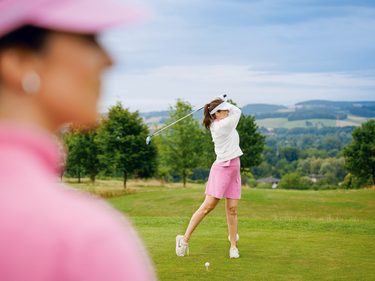 Golferin beim Abschlag auf einem Golfplatz, mit unscharfer Person im Vordergrund und Hügellandschaft im Hintergrund.