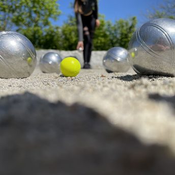 Metallkugeln und gelbe Zielkugel auf einem Kiesplatz, ein Gast spielt Boule im Hintergrund.