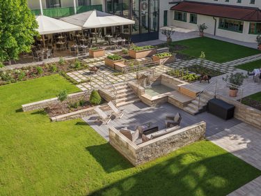 Blick auf einen Hotelinnenhof mit Terrasse, Sitzbereichen, Beeten, Treppen und großen Rasenflächen.