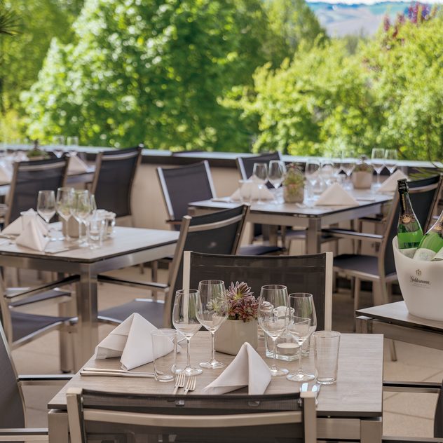 Gedeckte Tische auf einer Restaurantterrasse mit Gläsern, Servietten und Flaschenkühler vor grünen Bäumen.