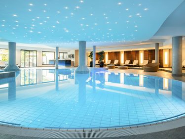 Großer Innenpool im Wellnessbereich mit Liegen, Säulen und beleuchteter Decke.