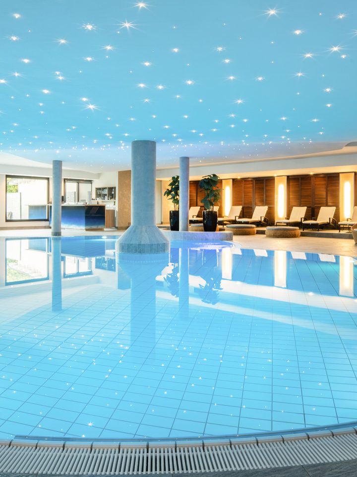 Großer Innenpool im Wellnessbereich mit Liegen, Säulen und beleuchteter Decke.