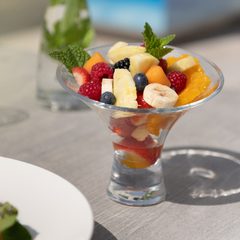 Bunter Obstsalat im Glasbecher auf einer sonnigen Terrasse, daneben ein Getränk mit Minze.
