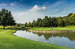 Golfer auf einem Grün neben einem Teich, umgeben von Rasen, Bäumen und offenem Himmel.