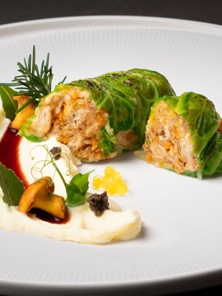 Aufgeschnittene Wirsingroulade mit Püree, Sauce und Garnitur auf einem weißen Teller.
