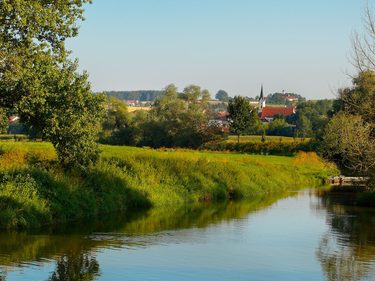 Ruhiger Fluss mit grünen Ufern, Bäumen und einem Dorf mit Kirchturm im Hintergrund.
