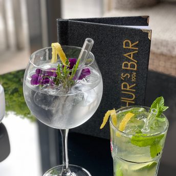 Zwei Cocktails mit Eis, Minze, Zitruszeste und Blüten stehen auf einem Tisch vor einer Barkarte.