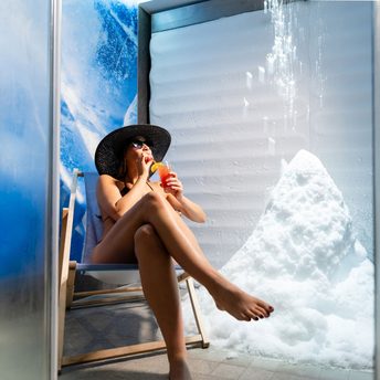 Eine Frau sitzt mit Cocktail und Sonnenhut auf einem Liegestuhl neben einem Schneehaufen in einem Wellnessraum.