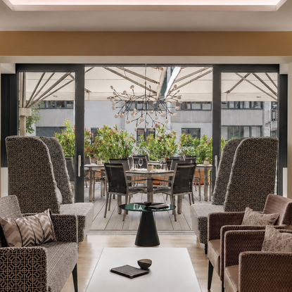 Sitzgruppe in einer Hotellobby vor großer Glasfront mit Blick auf eine möblierte Terrasse mit Pflanzen.