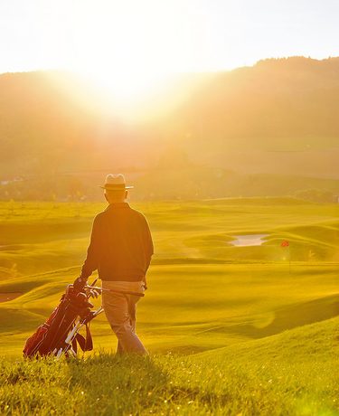 Ein Golfer mit Golftasche steht im Gegenlicht auf einem Golfplatz mit Hügeln und Bunkern bei Sonnenuntergang.