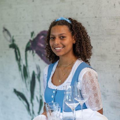 Eine Servicekraft in blauer Tracht trägt ein Tablett mit mehreren Gläsern in einem Innenraum.