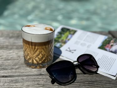 Glas mit Kaffee, Sonnenbrille und Magazin auf einem Holztisch am Pool.