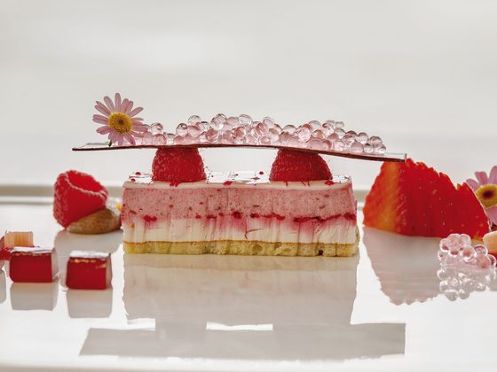 Ein geschichtetes Beerendessert mit Erdbeeren, Himbeeren, Geleewürfeln und Dekoration auf weißer Fläche.