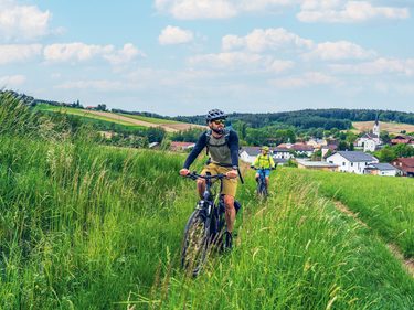 Zwei Radfahrer fahren auf einem schmalen Weg durch eine grüne Wiese vor einem Dorf mit Kirche.