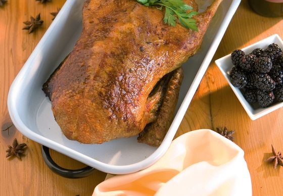 Gebratene Ente in einer Servierform mit Kräutern, Orangenscheibe, Sauce, Brombeeren und Rotwein auf Holztisch.