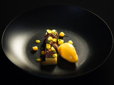 Modern angerichtetes Dessert mit Sorbet, Creme und Schokoladenelementen auf einem schwarzen Teller.