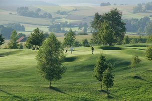 Drei Golfer auf einem Grün eines Golfplatzes in hügeliger Landschaft mit Bäumen und Feldern.