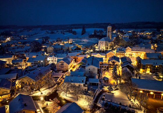 Luftaufnahme eines verschneiten Dorfes bei Nacht mit beleuchteten Häusern, Straßen und einer Kirche im Zentrum.