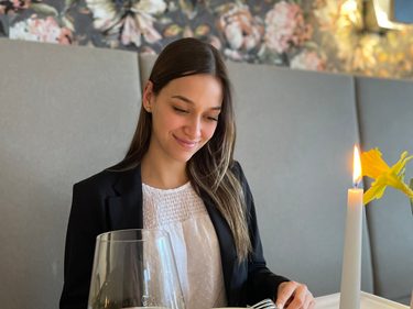 Eine Frau sitzt im Restaurant vor einer angerichteten Vorspeise mit einem Glas Weißwein und einer Kerze.