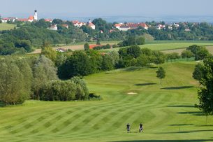Weitläufiger Golfplatz mit zwei Spielern auf dem Fairway, Bäumen und einem Dorf im Hintergrund.