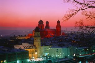 Verschneite Altstadt am Fluss mit beleuchteter Kirche und historischen Gebäuden bei rosa Abendhimmel.