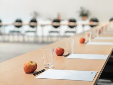 Lange Tischreihe in einem Seminarraum mit Gläsern, Schreibunterlagen, Stiften und Äpfeln.