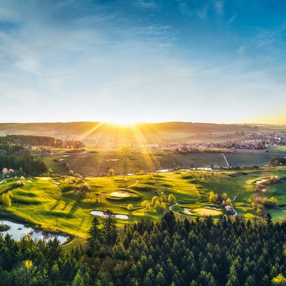 Panoramablick auf einen Golfplatz mit Hügeln, Teichen, Wald und tief stehender Sonne.