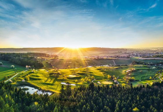 Panoramablick auf einen Golfplatz mit Hügeln, Teichen, Wald und tief stehender Sonne.