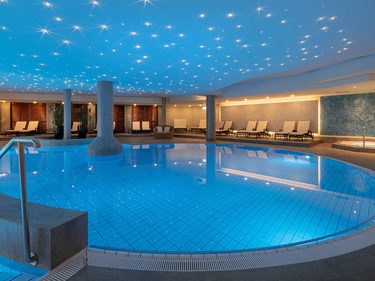 Runder Innenpool im Wellnessbereich mit beleuchteter Decke, Liegen und Whirlpool.