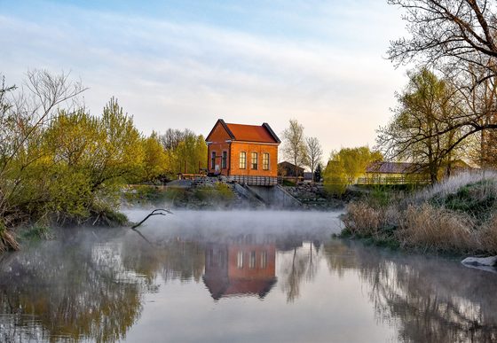 Kleines Haus am Ufer eines ruhigen Gewässers mit Spiegelung im Wasser und leichtem Morgennebel.