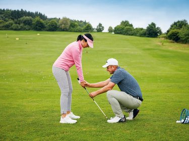 Ein Golflehrer unterstützt eine Spielerin beim Training mit Golfschläger auf einer grünen Übungsfläche.