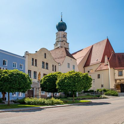Historische Kirche mit Zwiebelturm an einem sonnigen Ortsplatz, flankiert von Bäumen und Gebäuden.