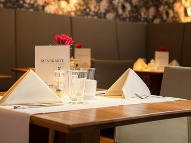Gedeckter Tisch in einem Restaurant mit Menükarte, Gläsern, Servietten und floraler Tapete im Hintergrund.