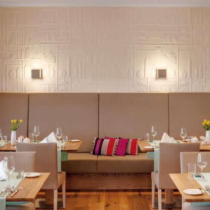 Restaurantbereich mit gedeckten Tischen, gepolsterter Sitzbank, Stühlen und Blumenvasen an einer dekorativen Wand.