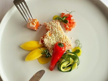 Angerichtetes Fischfilet mit Perlenkruste, Gemüse und Garnitur auf einem weißen Teller mit Besteck.