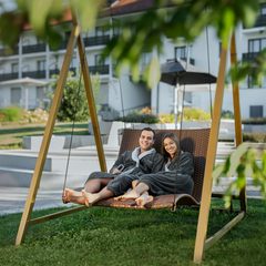 Ein Paar im Bademantel entspannt auf einer Hängeliege im Hotelgarten vor dem Gebäude.